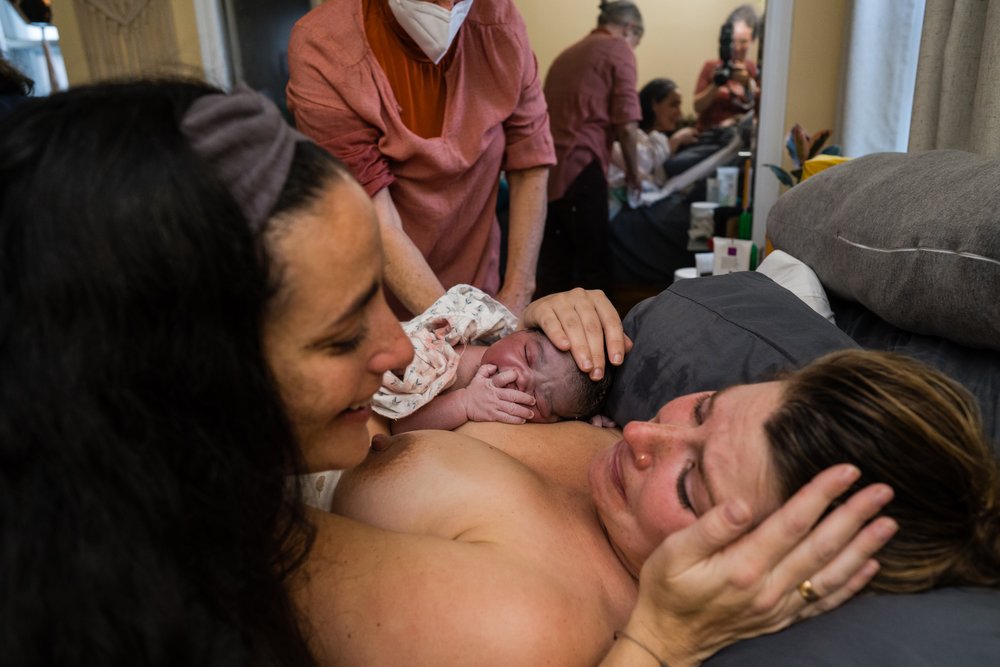 Doula caresses mom's face and smiles after she gave birth, baby skin to skin minutes old, midwife and photographer in background