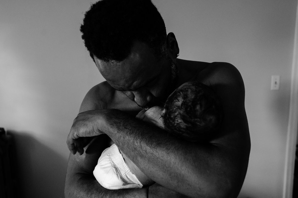 Black father wraps his new baby in his arms, skin to skin, kisses his new son, black and white portrait
