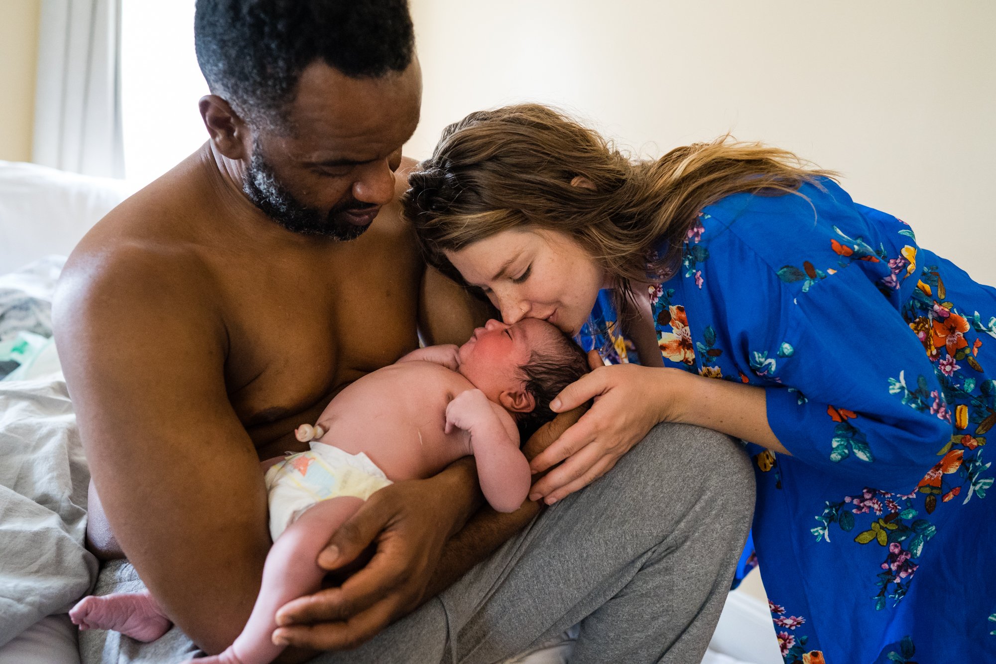 White mother in blue floral robe gently kisses newborn baby on the head while Black father holds him skin to skin, Philadelphia home birth photography