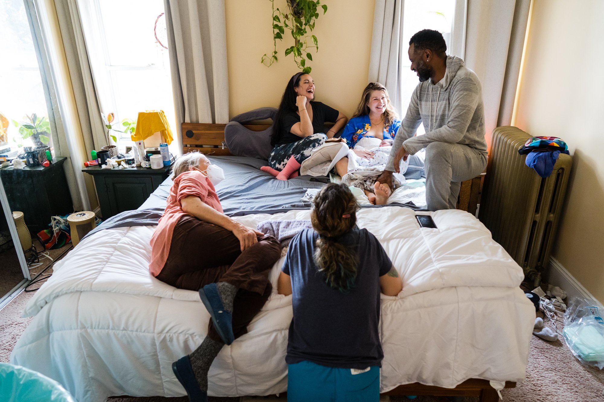 Home birth team relaxes on bed together, midwives, doula, father, laugh at story mom is telling, Philadelphia photographer