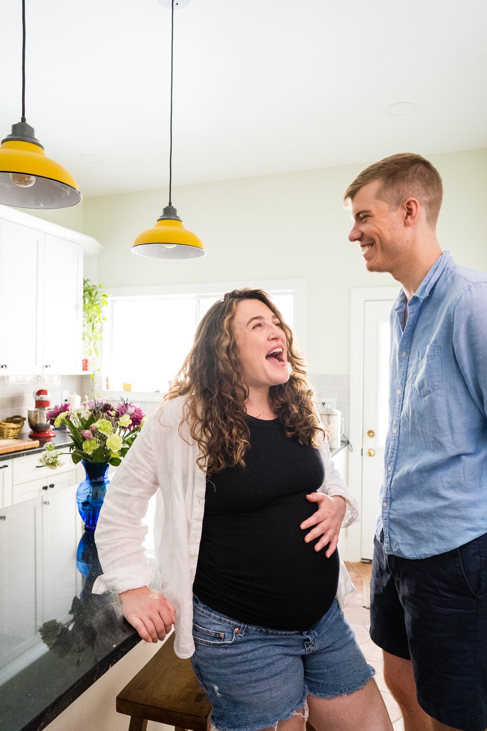 Pregnant mom laughs and holds her belly, dad smiles, bright colorful kitchen, at-home maternity session