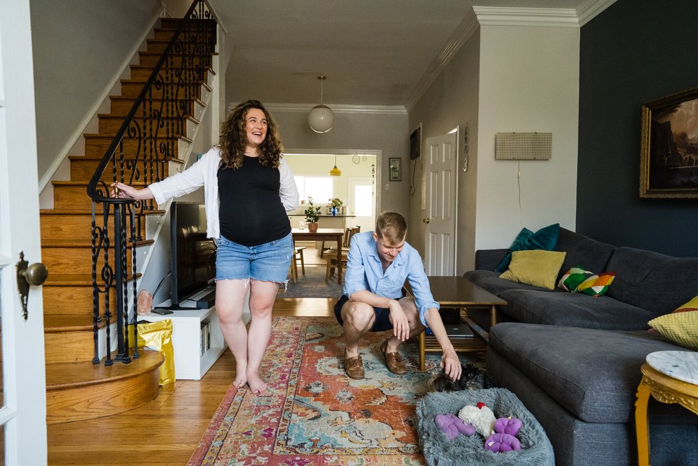 Couple stands in their South Philly home, dad pets dog while mom laughs and leans on the stairs, Philly maternity photos