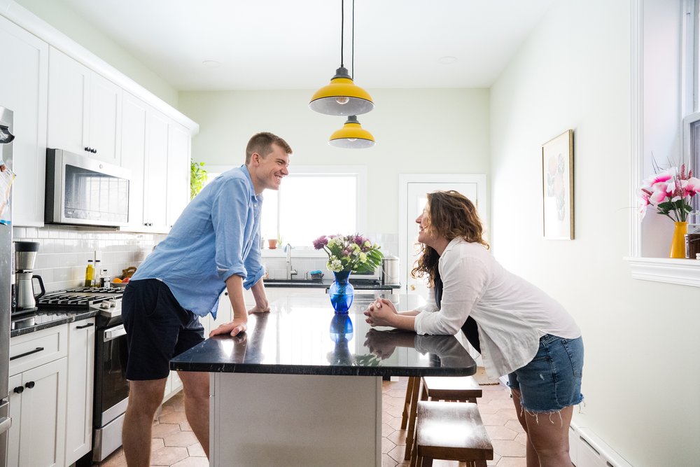 Couple leans over kitchen island, bright window light, smiles at each other, pregnant mom, Philadelphia maternity photographer