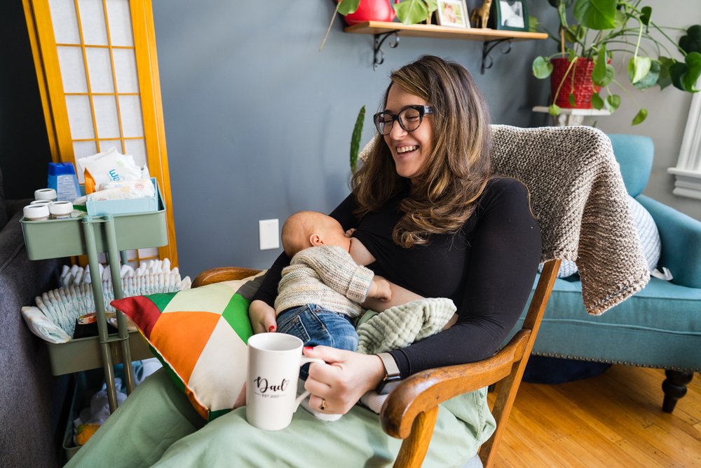 New mom breastfeeds in a rocking chair, smiles, drinks coffee out of "dad" mug, Philadelphia newborn photography