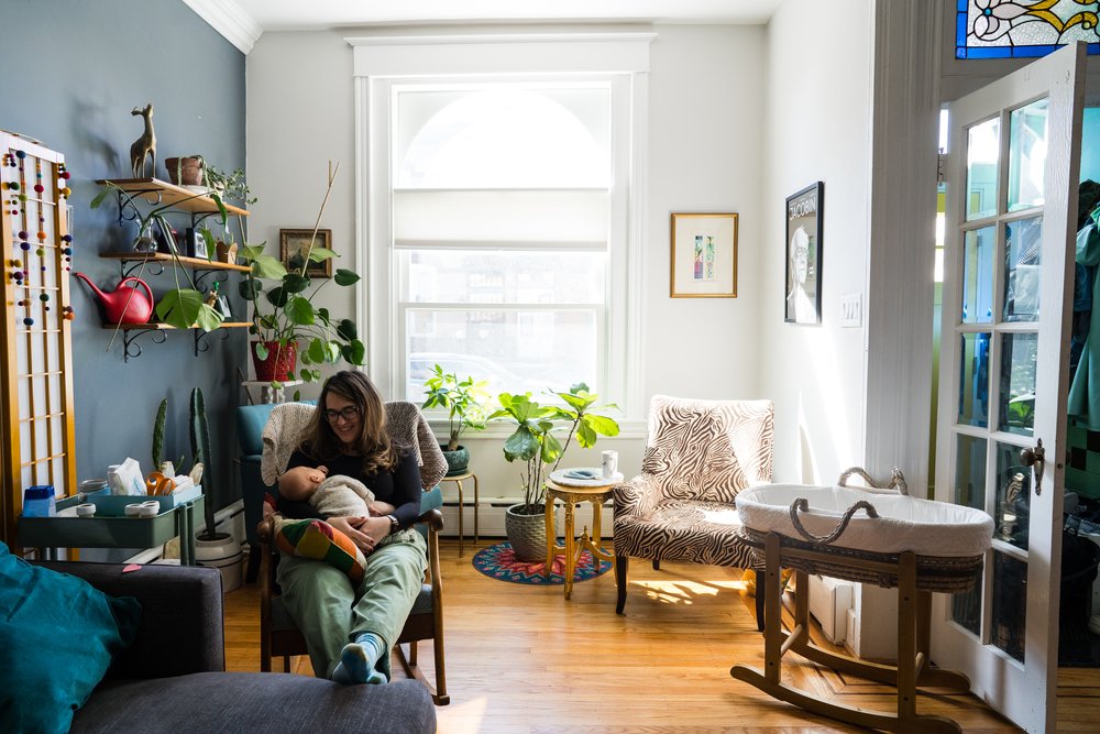 Mom sits in sunbathed south philly home, plants and cute decor, breastfeeding baby in rocking chair and smiling, Philadelphia photographer