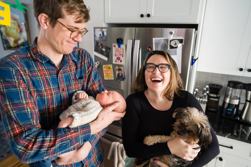 Dad holds newborn and smiles, mom holds dog like baby and laughs, Philadelphia in-home photography