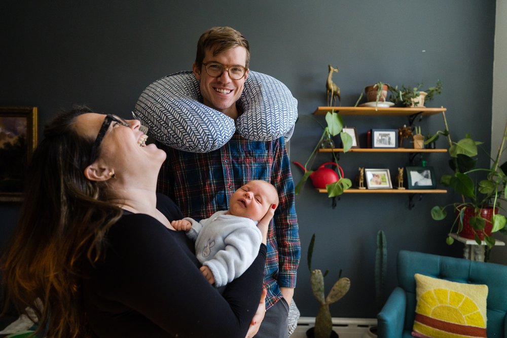Mom holds baby and has a huge laugh at dad wearing nursing pillow around his neck, Philadelphia family photography