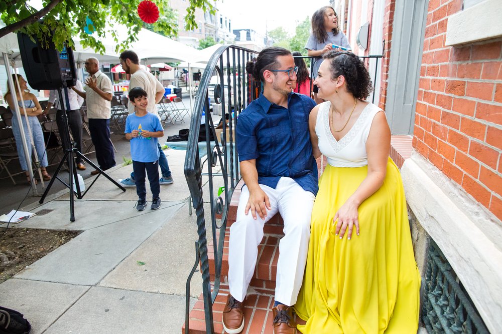 Bride and groom sit on the stoop of childhood rowhome as neighbors get ready for block party wedding, Philadelphia documentary photographer