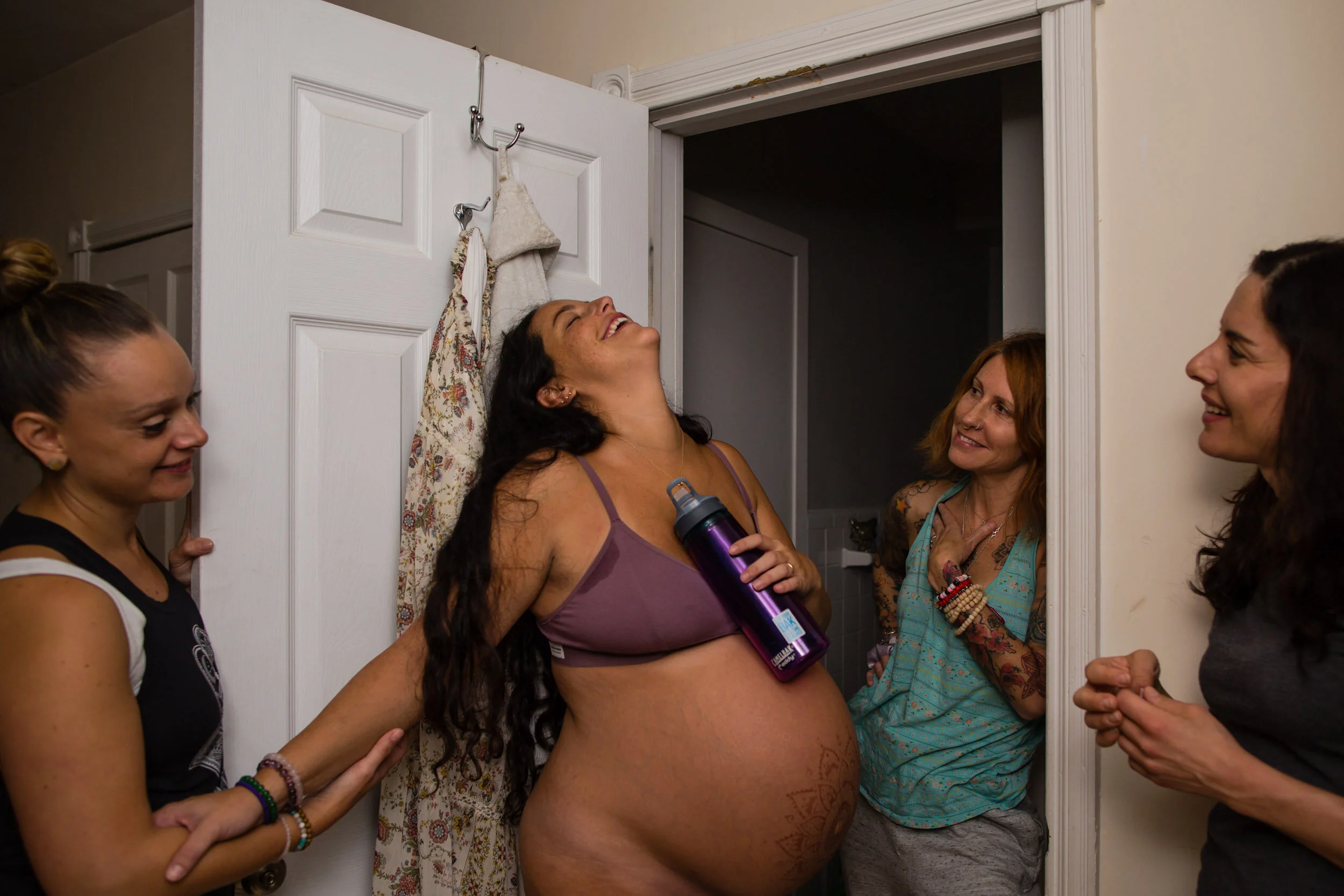 Laboring woman throws her head back to laugh in between contractions surrounded by her doulas and friends, Philadelphia birth photographer