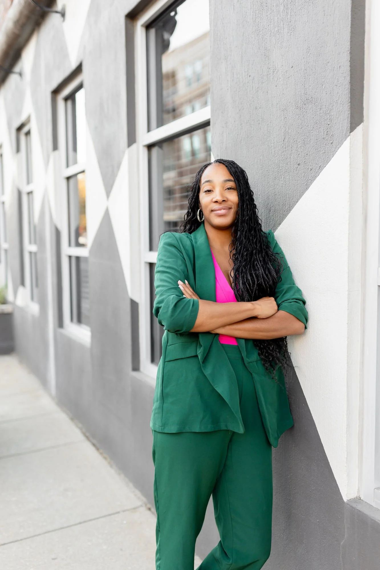 A woman with long curly black hair wearing a green blazer and pants, pink shirt, standing with arms crossed against a wall with gray, white, and black geometric patterns, outdoors in daylight.