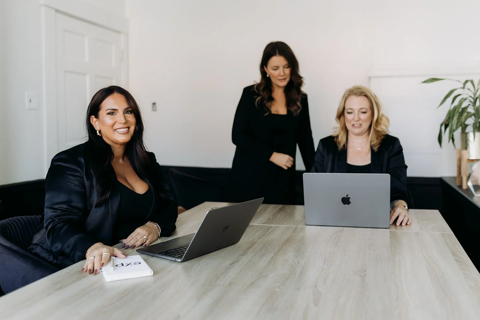Three women in a business meeting; two seated at a conference table with laptops, one standing between them.