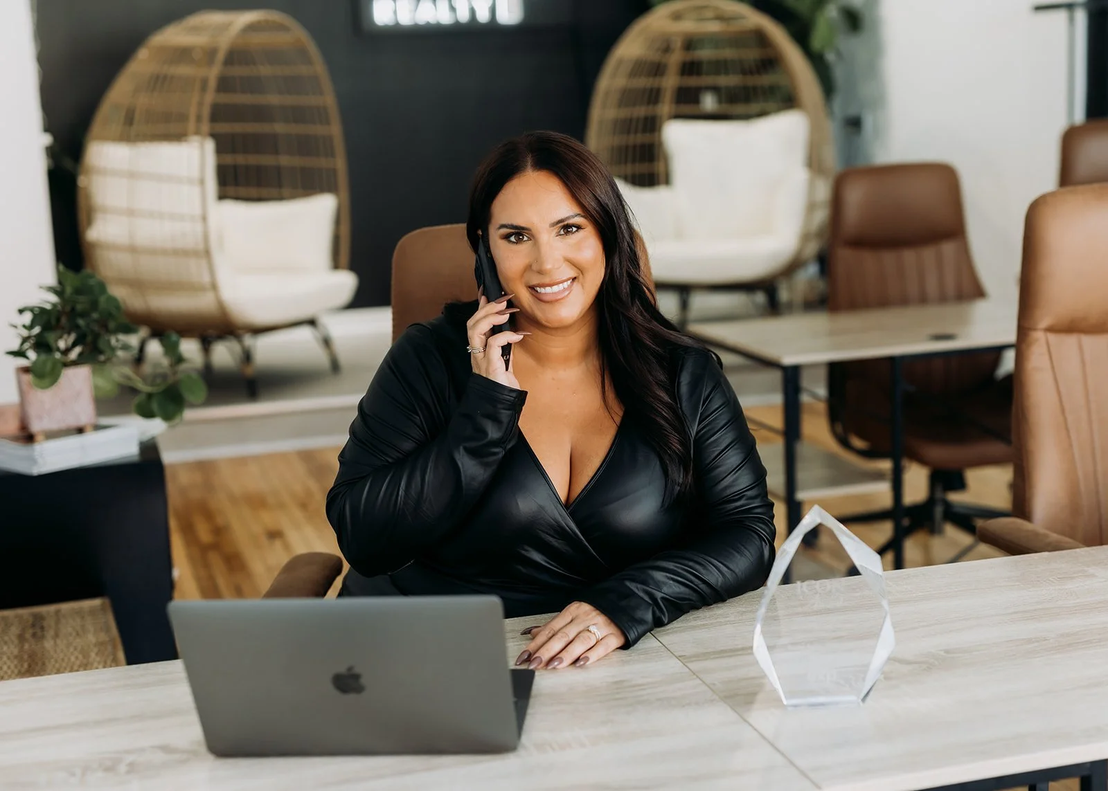 A woman with long dark hair, wearing a black leather jacket, sitting at a desk with a laptop, talking on the phone in a modern office with hanging chairs and a plant in the background.