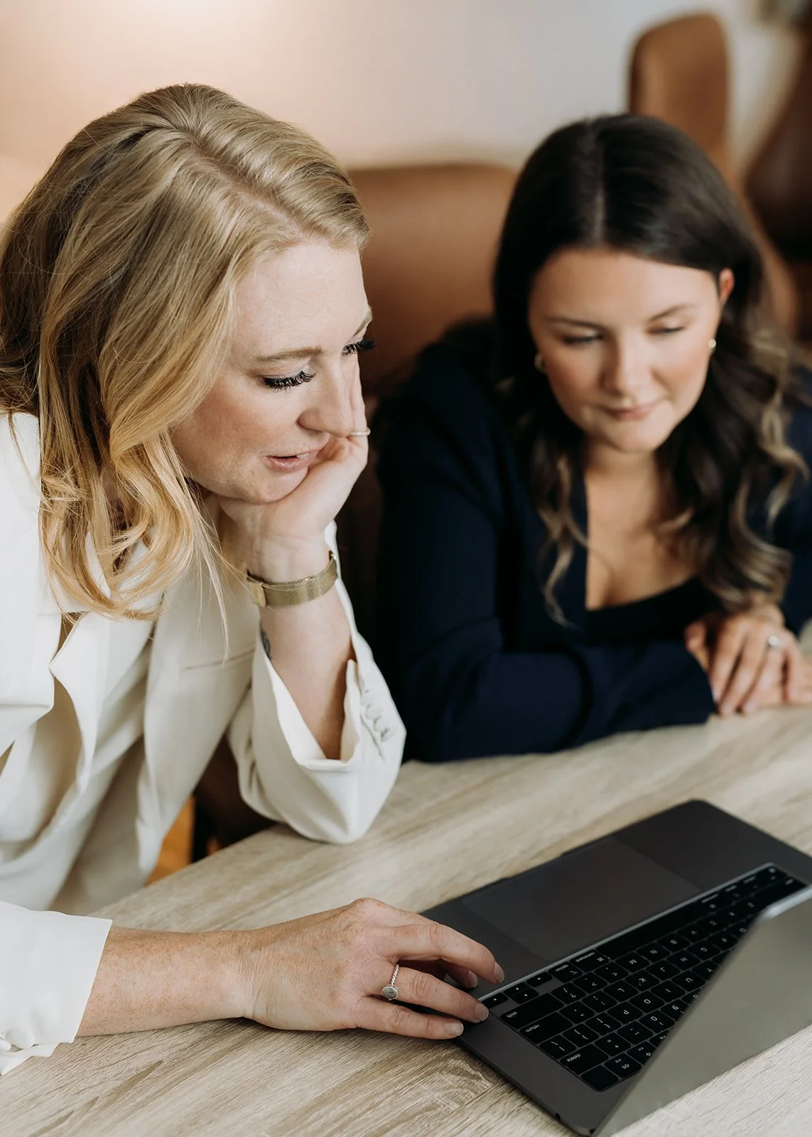 Two women sit at a wooden table looking at a laptop screen, engaged in conversation.