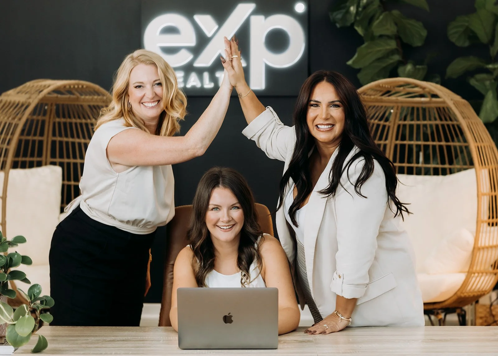 Three women celebrating with a high five in an office setting, with a laptop on the table and a sign reading 'expo' in the background.