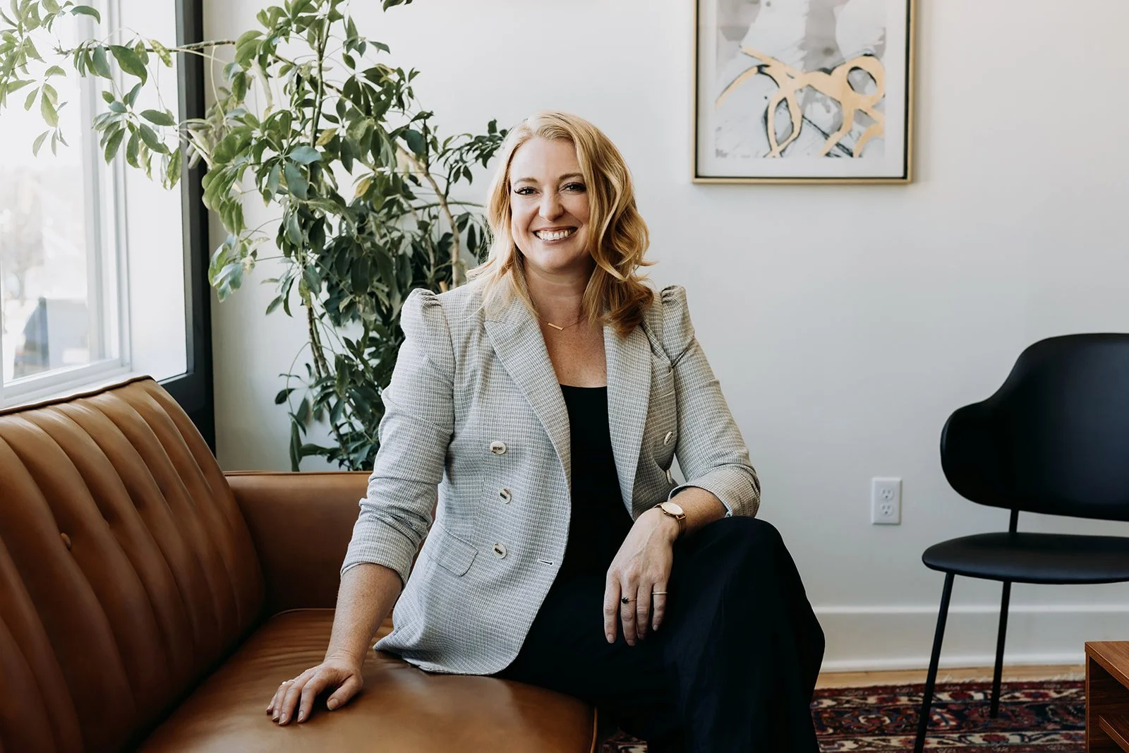 A woman with shoulder-length blonde hair, wearing a dark blazer and white top, sitting at a desk with a laptop in front of her, smiling at the camera in a professional office setting.