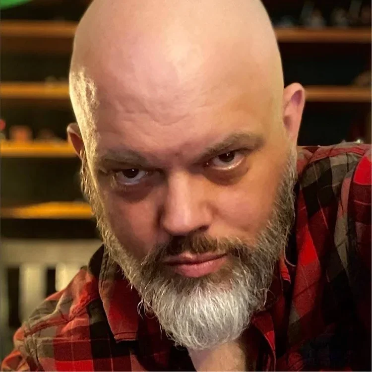 Smiling man with a beard wearing a black collared shirt standing outdoors with a large tree and green foliage in the background.
