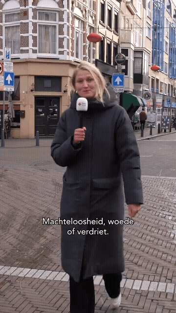 A woman holding a microphone standing on a city street with buildings, shop signs, and street signs in the background.