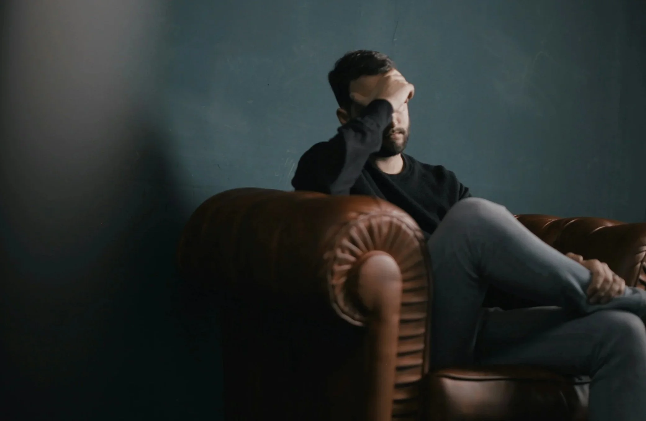 A person wearing a black shirt, sitting on a brown leather couch against a plain, dark-colored wall, resting their head in their hand.