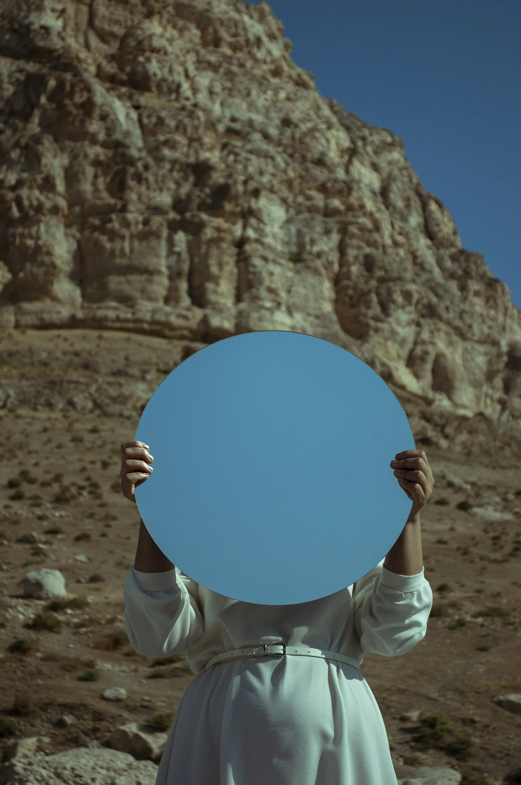 Person whose face cannot be seen, holding a large blue circled mirror in front of their face, standing in a desert landscape with rocky hills under a blue sky.
