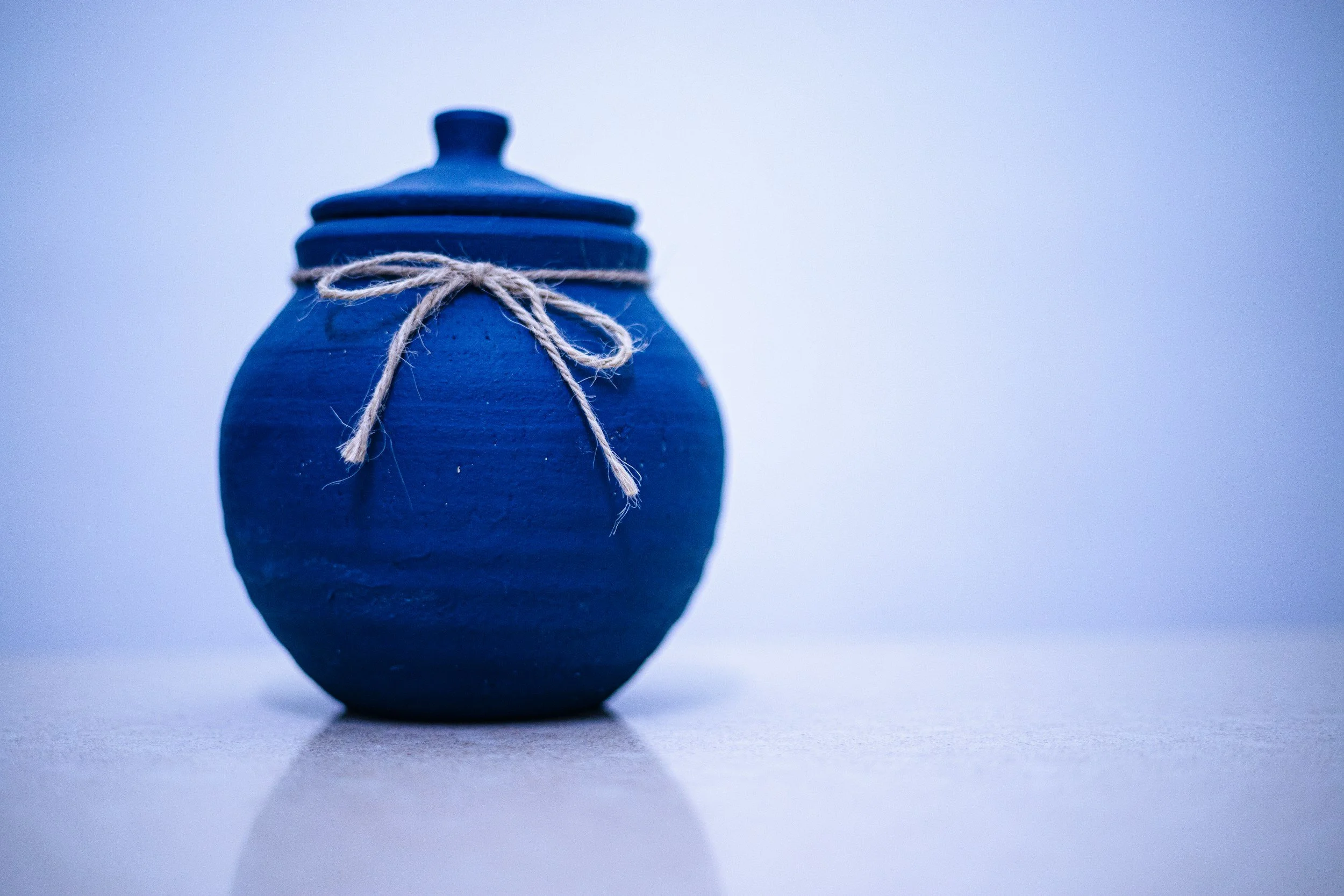 Blue ceramic vase with a narrow neck and round body, tied with a piece of twine, placed on a surface with a reflective shadow.