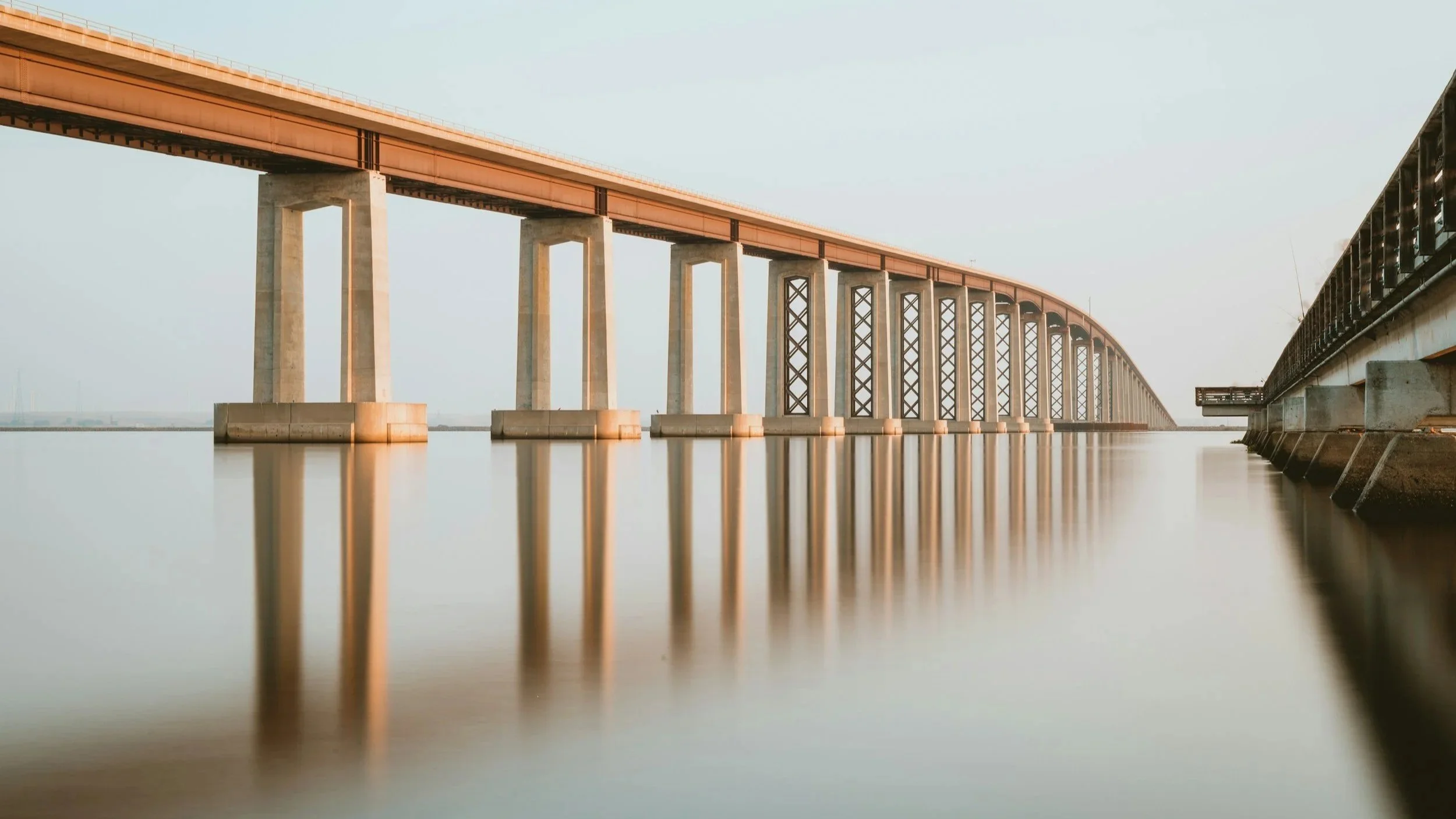 A long bridge extends over a calm body of water, supported by large concrete pillars, with a clear sky overhead.