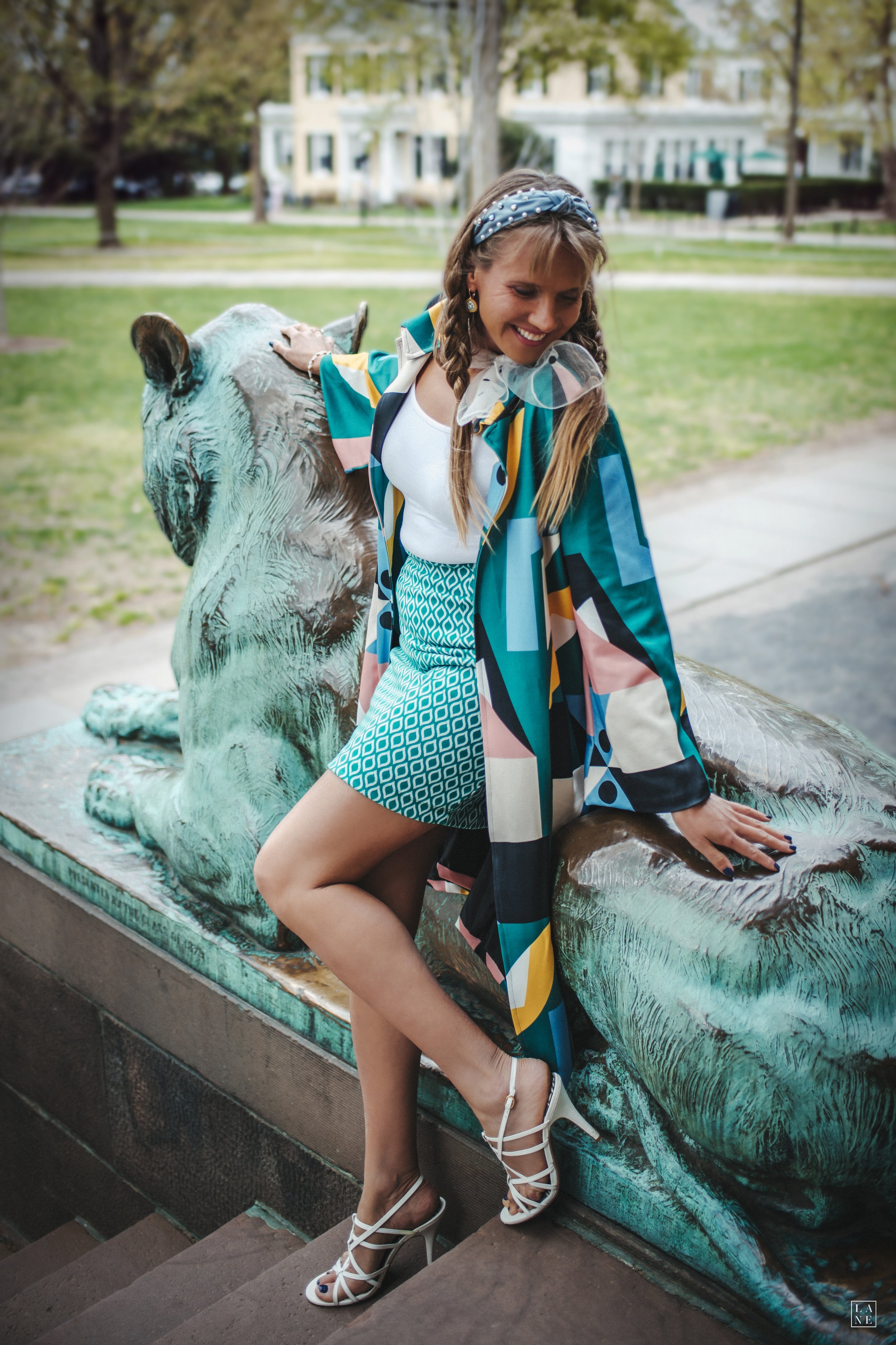 Woman sitting on a tiger statue, wearing a colorful coat, patterned skirt, and white sandals outdoors.