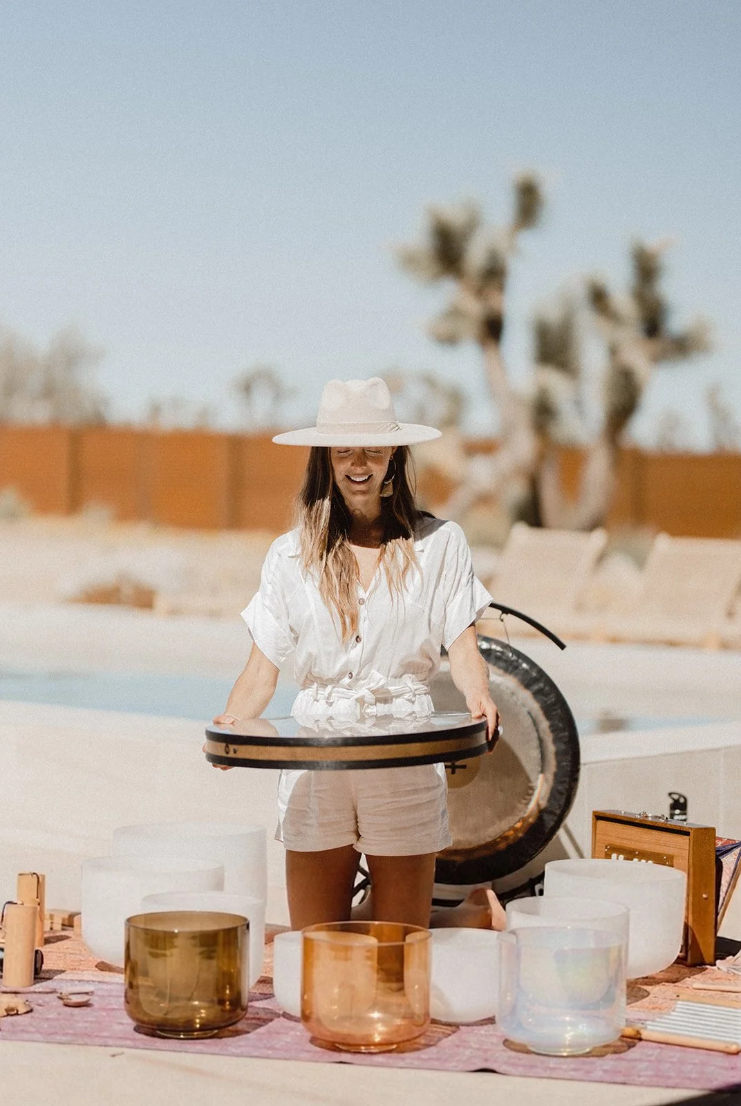 A woman wearing a white summer outfit and wide-brimmed hat, smiling while holding an Ocean drum, with other crystal bowls around her and desert landscape with a cactus in the background.