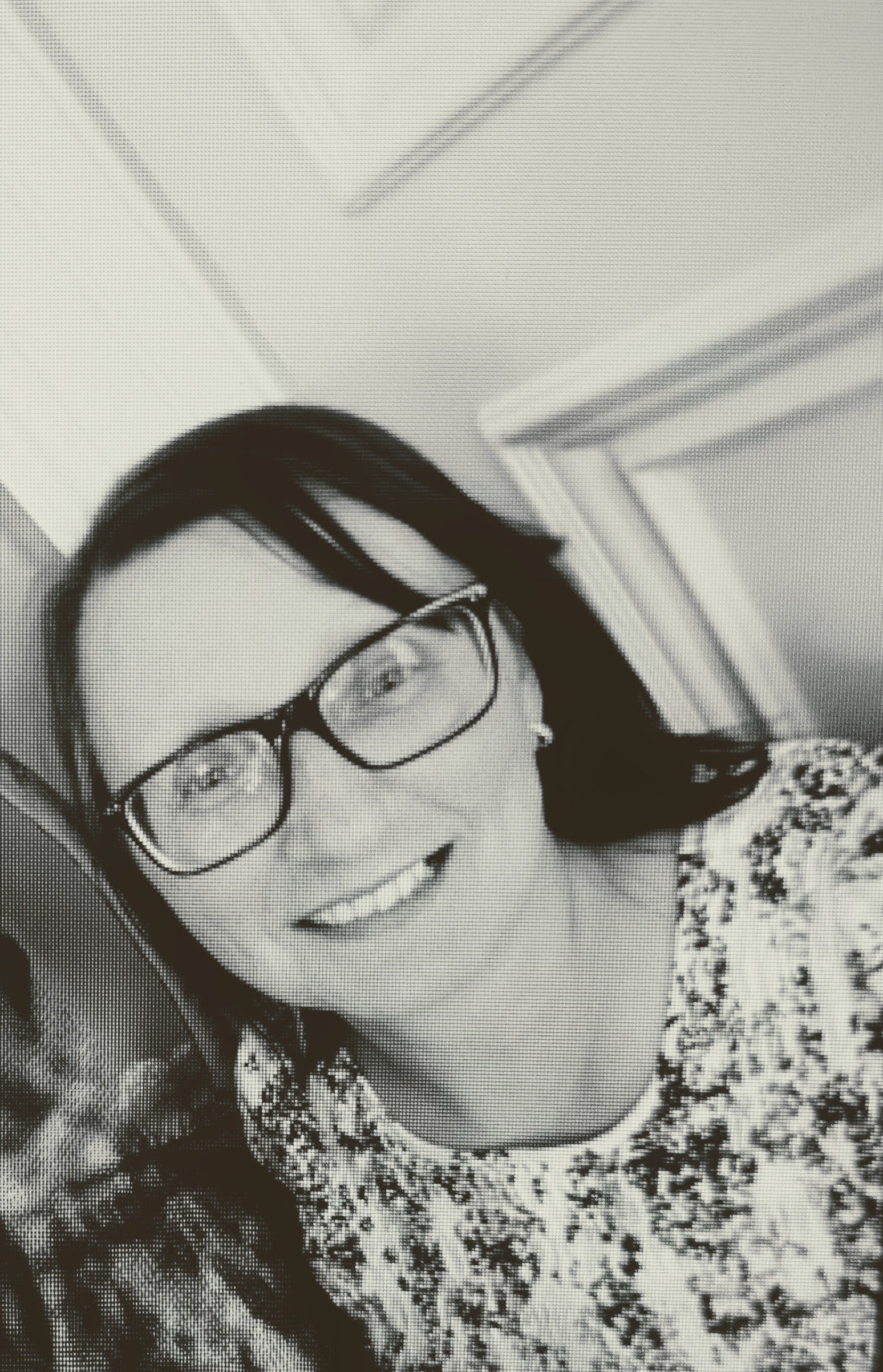 A black and white photo of a woman with shoulder-length dark hair, wearing glasses and a patterned shirt. She is smiling and looking slightly to her left, positioned indoors near a doorway or wall.