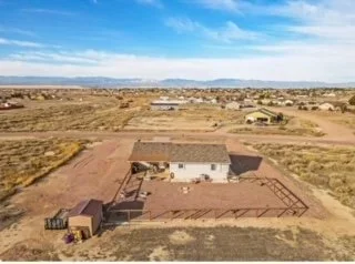 A rural property with a house, fenced yard, and a small shed, situated in a desert landscape with open fields and mountains in the background.