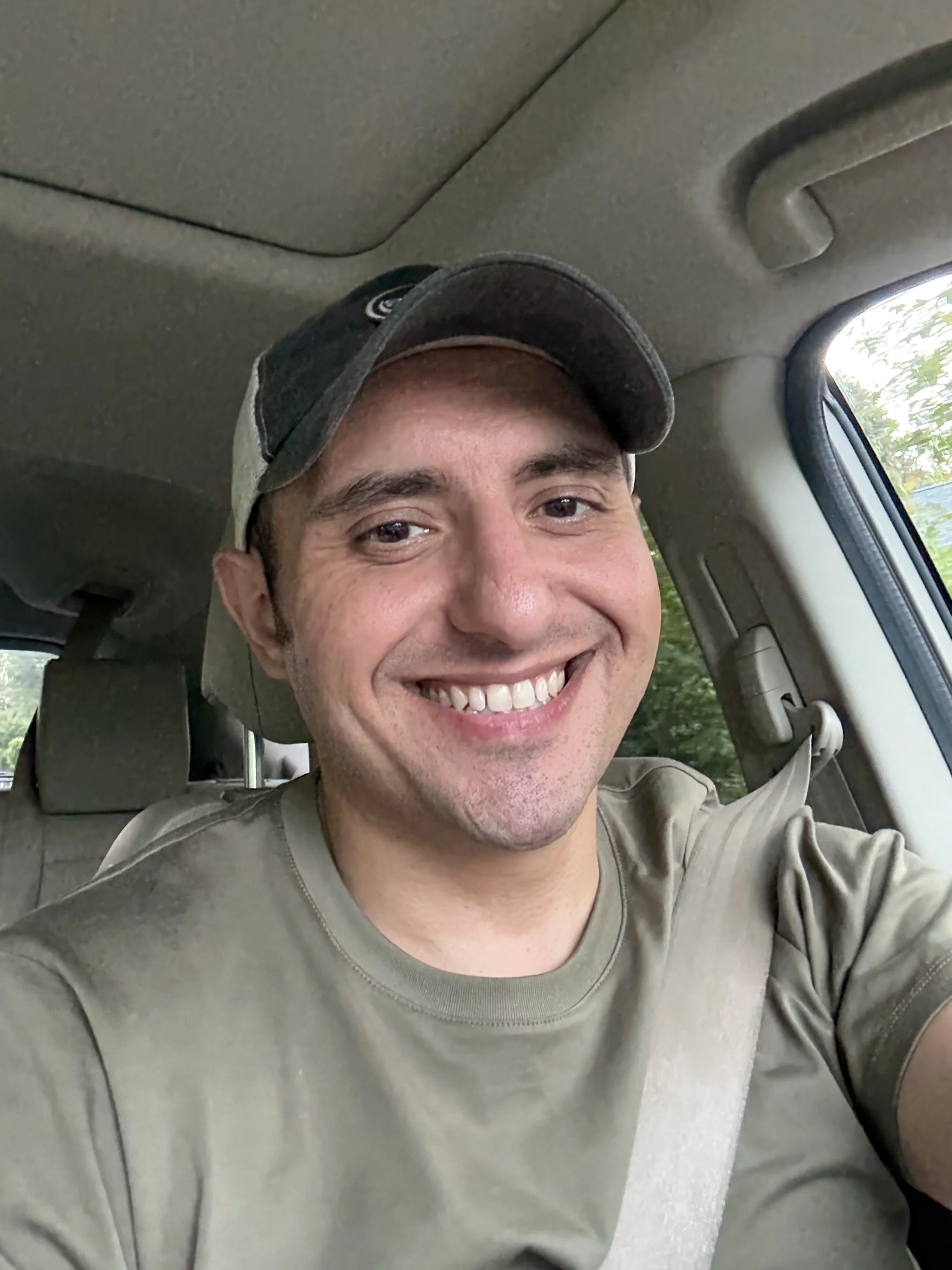 A smiling man wearing a khaki shirt and a baseball cap sitting in a vehicle, with a seatbelt across his chest. The background shows the car interior and a window with trees outside.