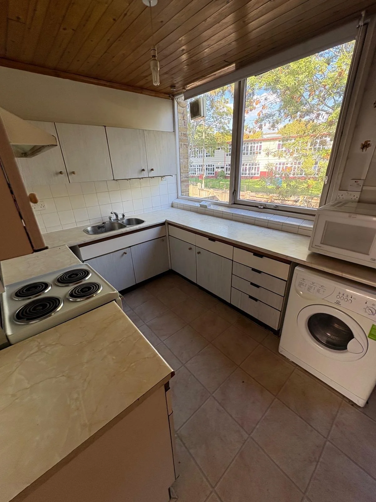 Kitchen with wooden ceiling, white cabinets, electric stove, double sink, microwave, washing machine, large window showing trees and a building outside.