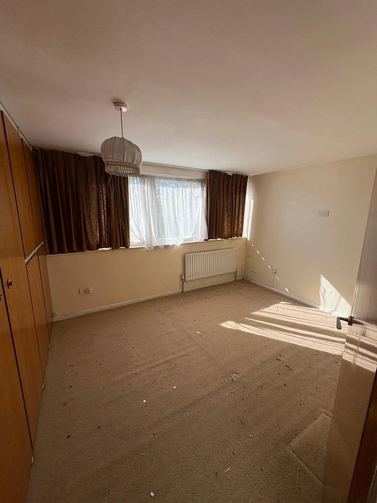 Empty room with beige carpet, cream-colored walls, and a large window with brown curtains and sheer white curtains, sunlight streaming in.