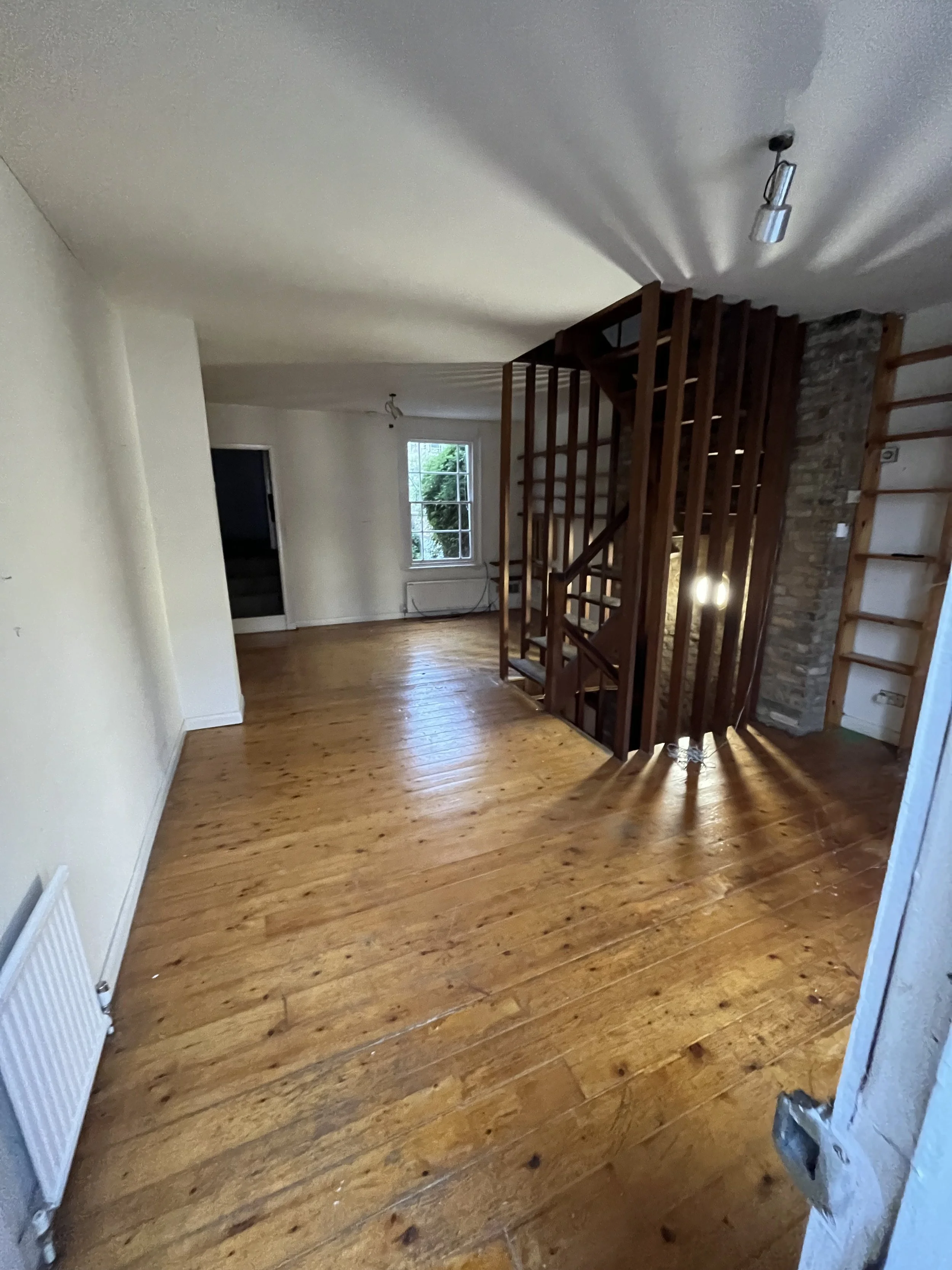 Empty living room with wooden floors, white walls, and a staircase with wooden railing leading to the upper floor. There is a window and a door at the far end of the room. A radiator is on the left wall.