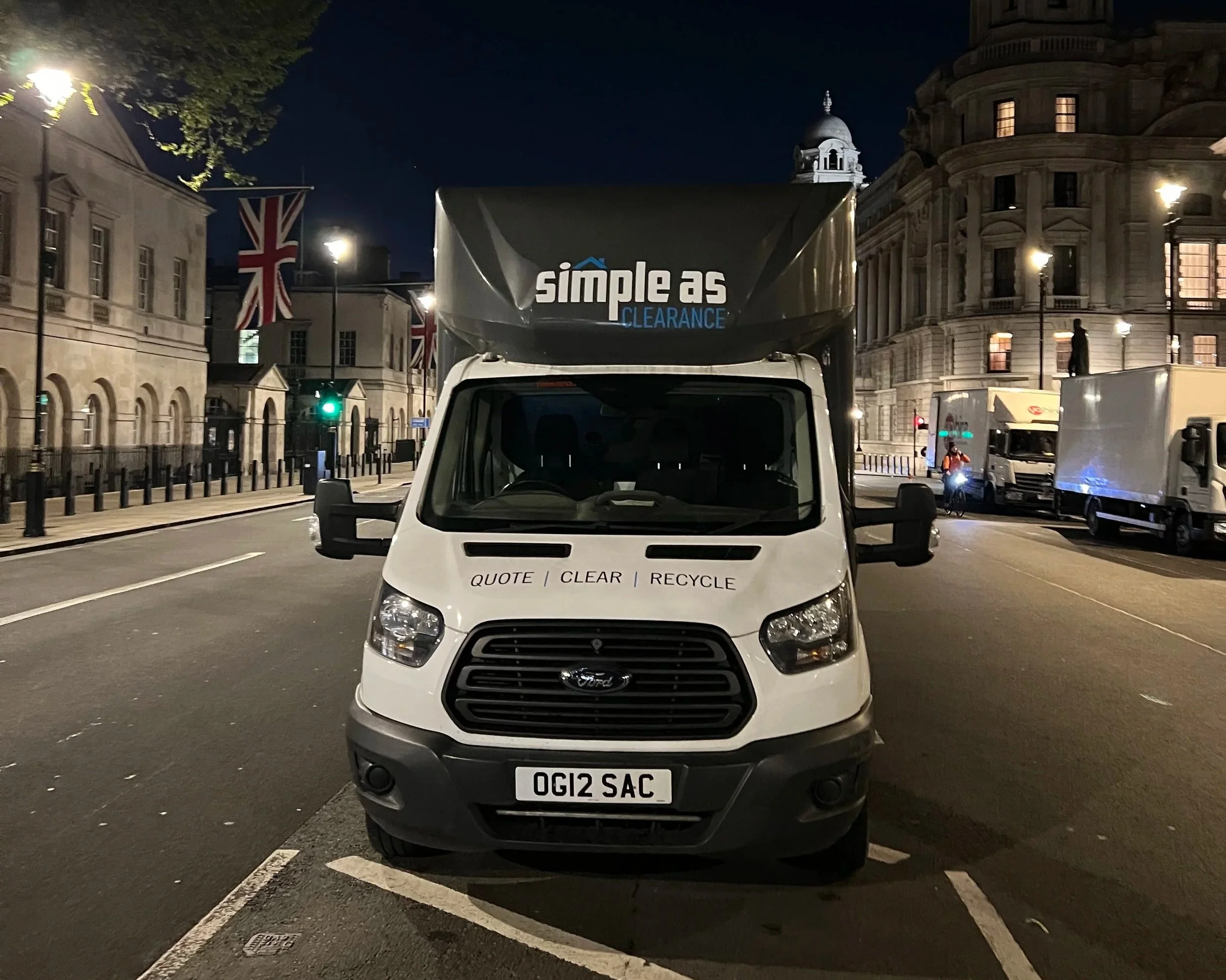 White Ford truck parked on a city street at night, with buildings and lampposts illuminated in the background, and a Union Jack flag hanging on a building.