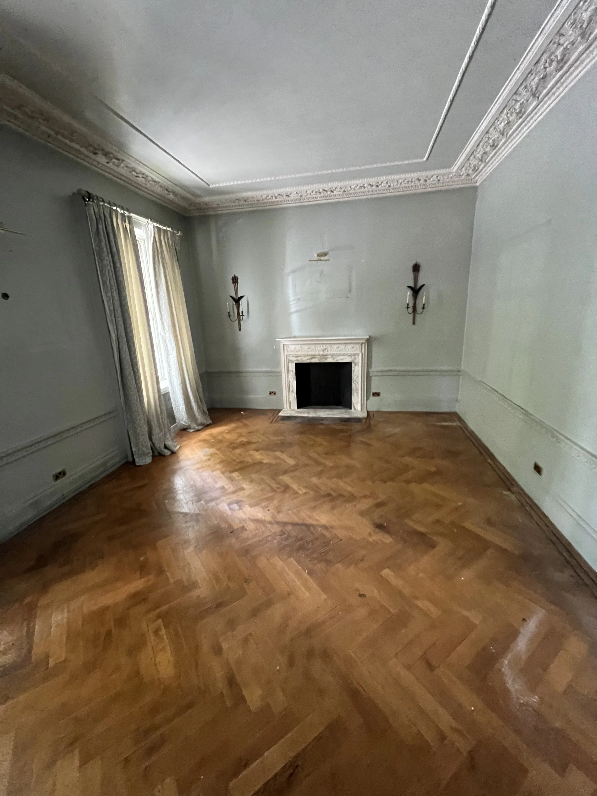 Empty room with hardwood floor, two wall-mounted candle sconces, a fireplace, and curtains on a window, with the ceiling detailed with crown molding.