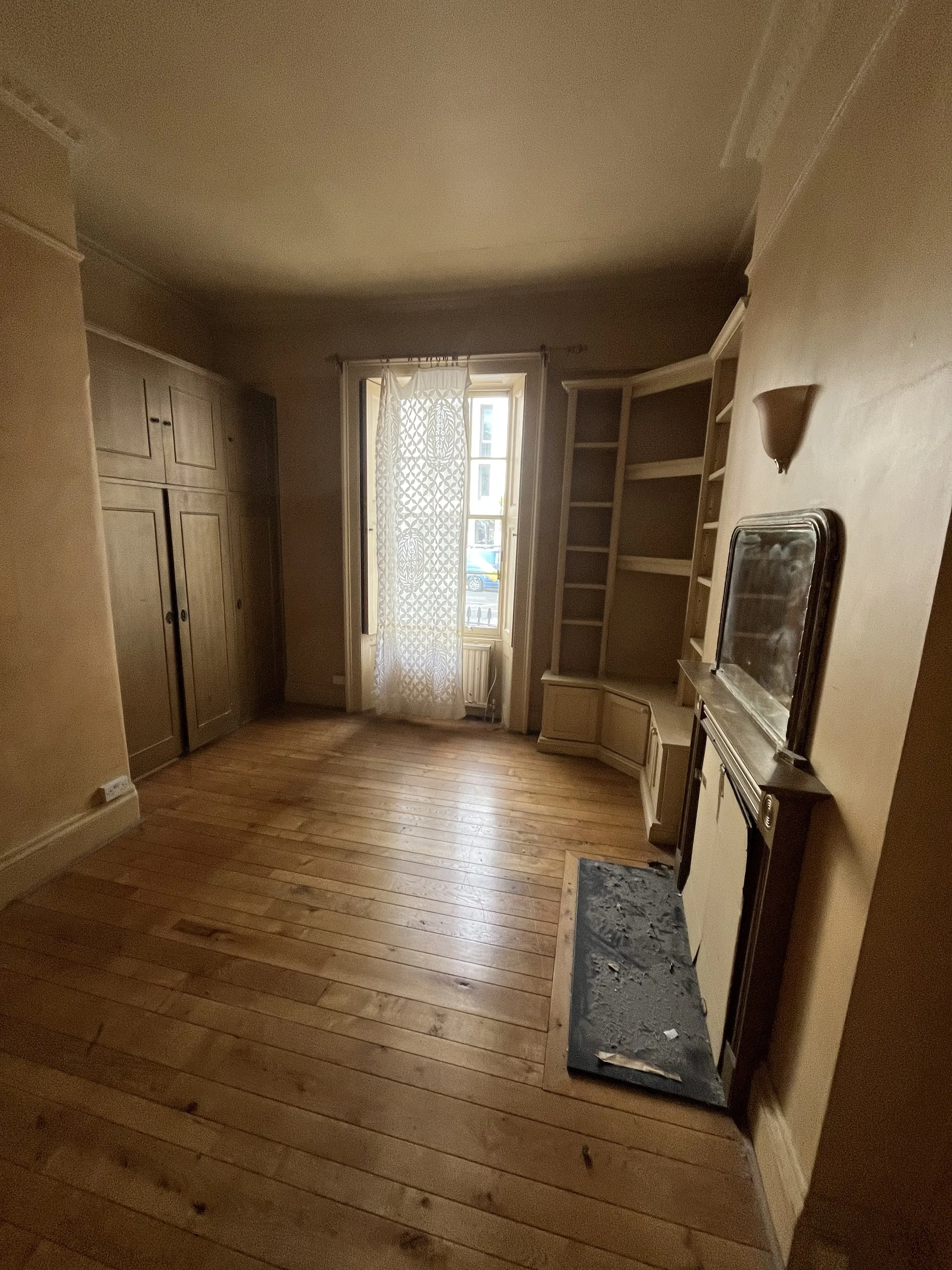 Empty living room with hardwood floors, a window with lace curtain, built-in shelves, a vintage mirror, and an old fireplace.