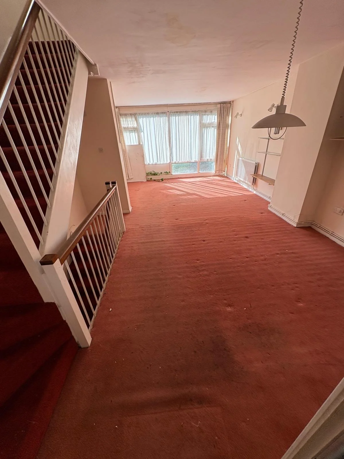 Empty living room with worn red carpet, peeling paint on ceiling, large window with curtains, staircase to the left, and a hanging lamp.
