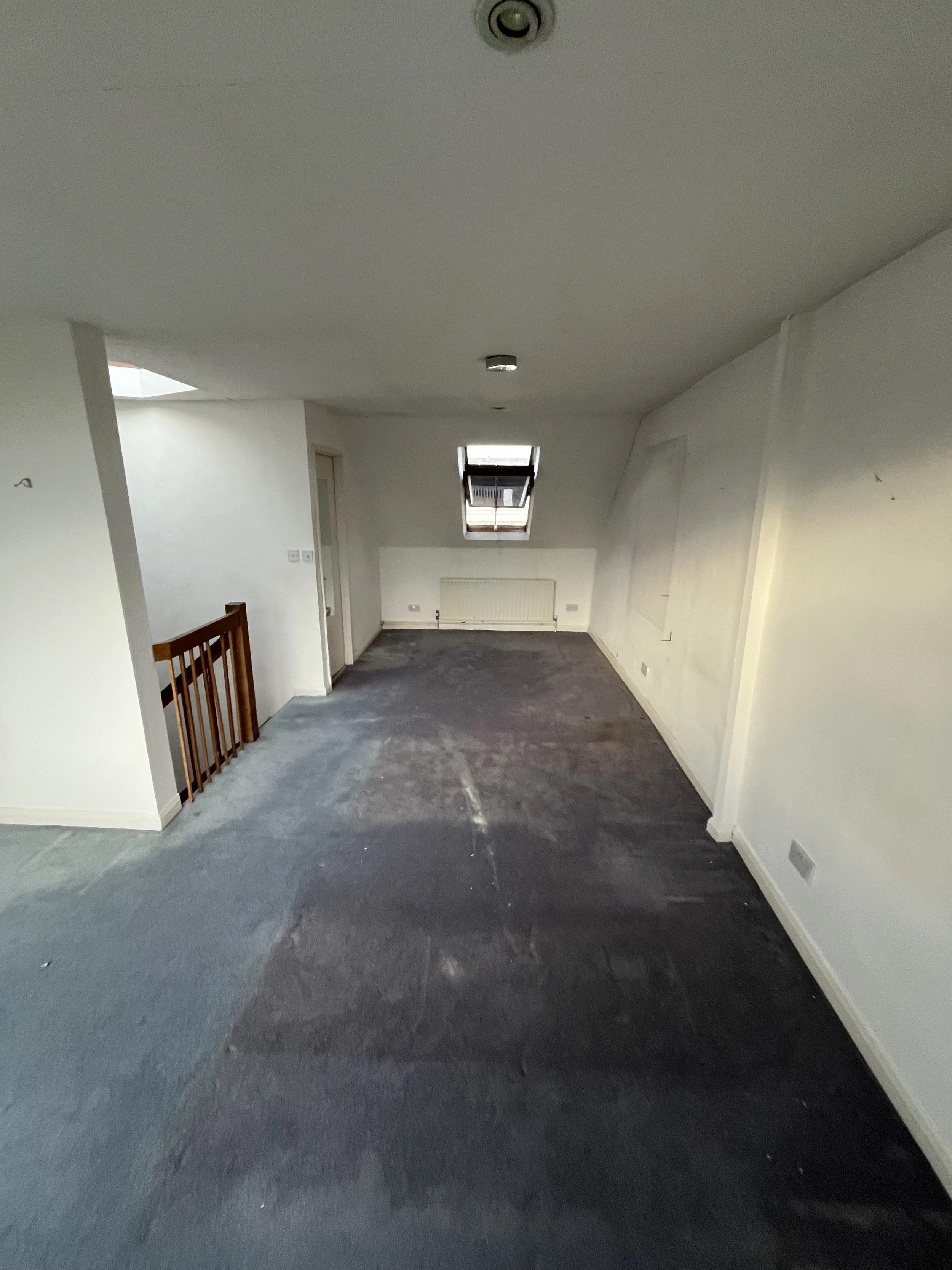 Empty room with slanted ceiling, dark carpet, small window, radiator, and white walls.