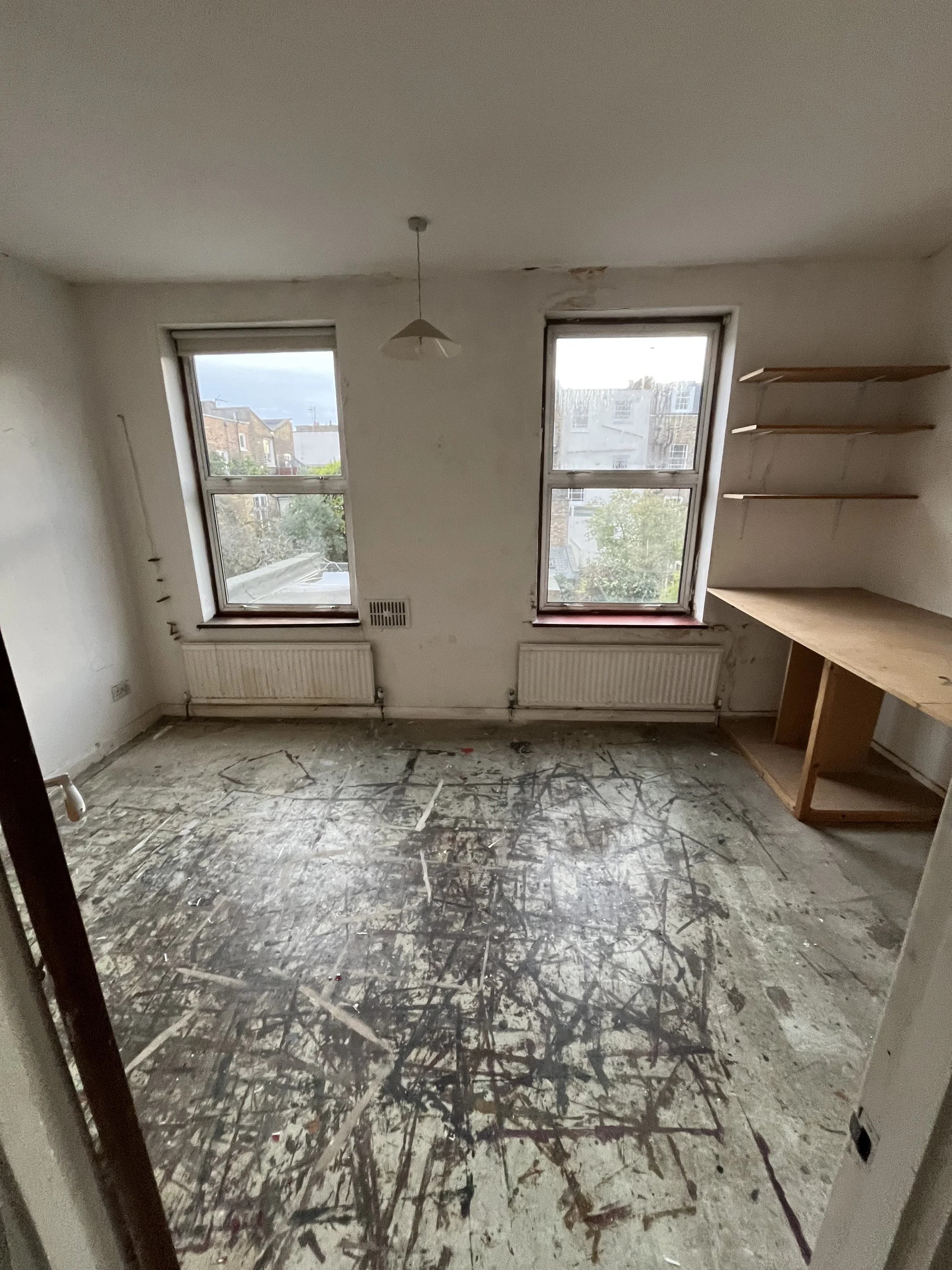 Empty room with two large windows, a dropped ceiling with a light fixture, a built-in radiator under each window, and a wooden shelves and work surface on the right wall. The floor is unfinished and marked with dark paint splatters.