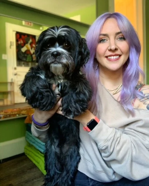 A young woman with pastel purple hair smiling while holding a small black and white dog indoors.