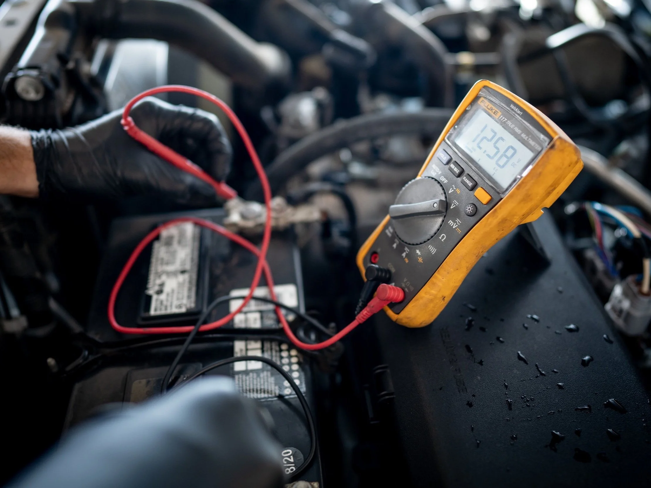 Photo showing handheld multimeter testing battery leads in engine bay.