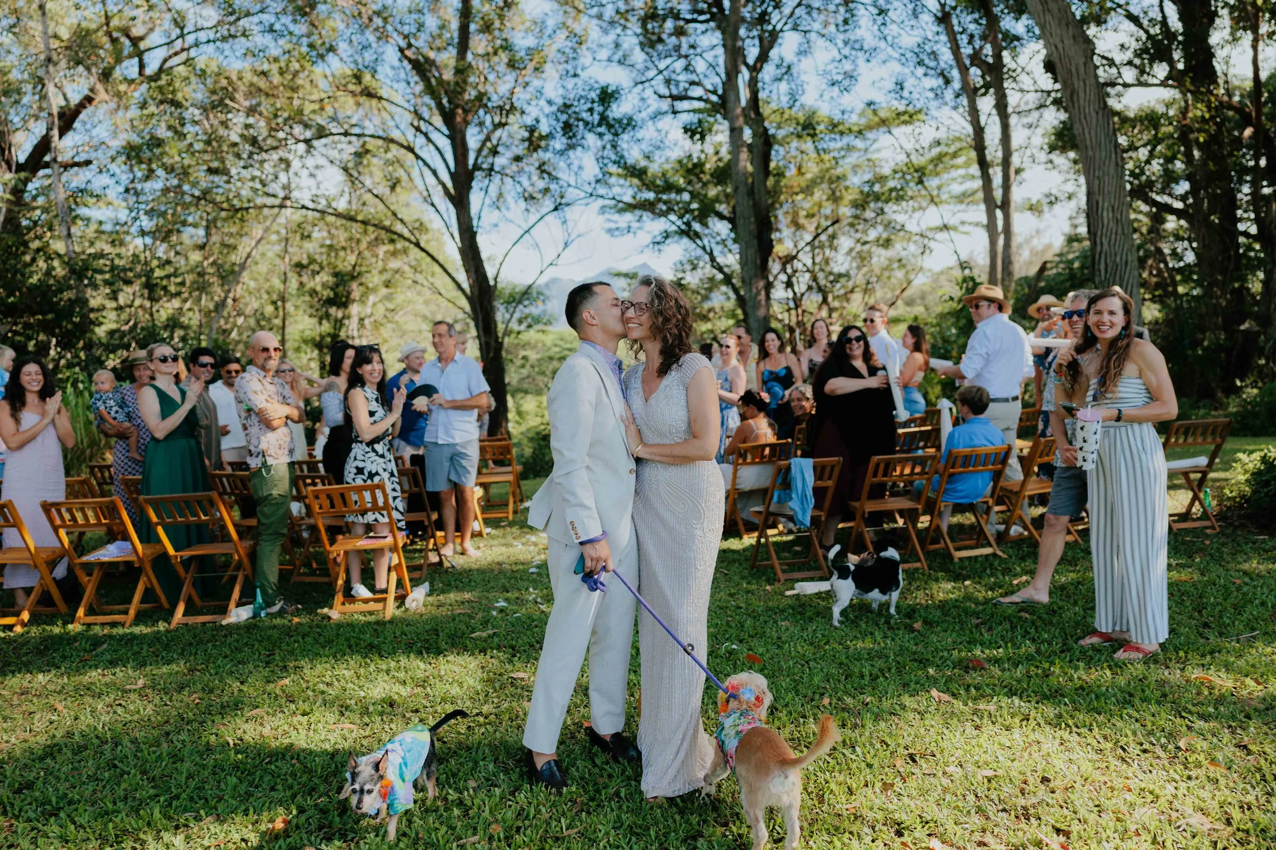 Couple in wedding attire sharing a kiss, surrounded by seated and standing guests, with dogs on leashes, outdoors in a park-like setting with trees and clear sky.