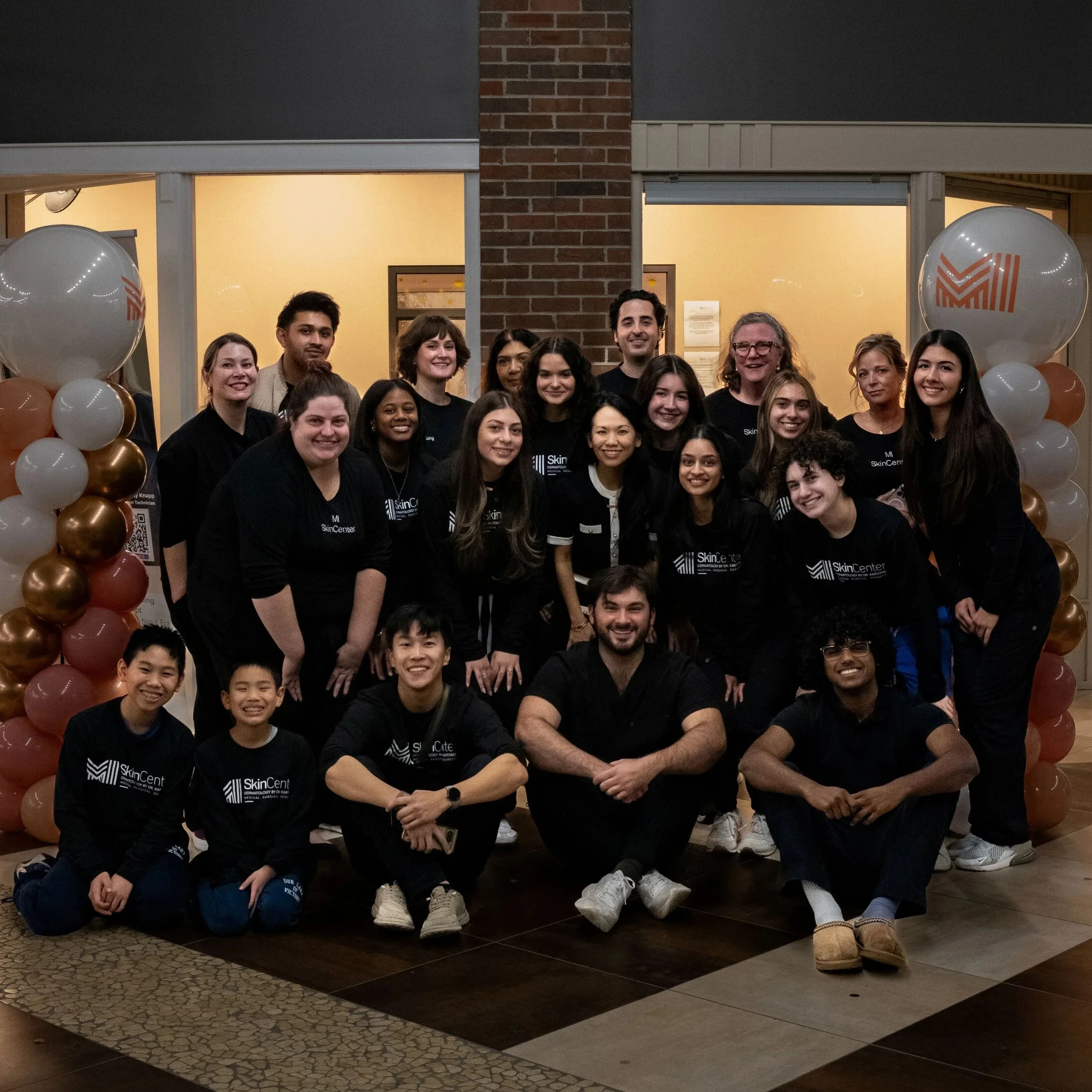Staff at MI Skin Center smiling, including children, posing indoors in a dermatology office with balloons and medical posters in the background.
