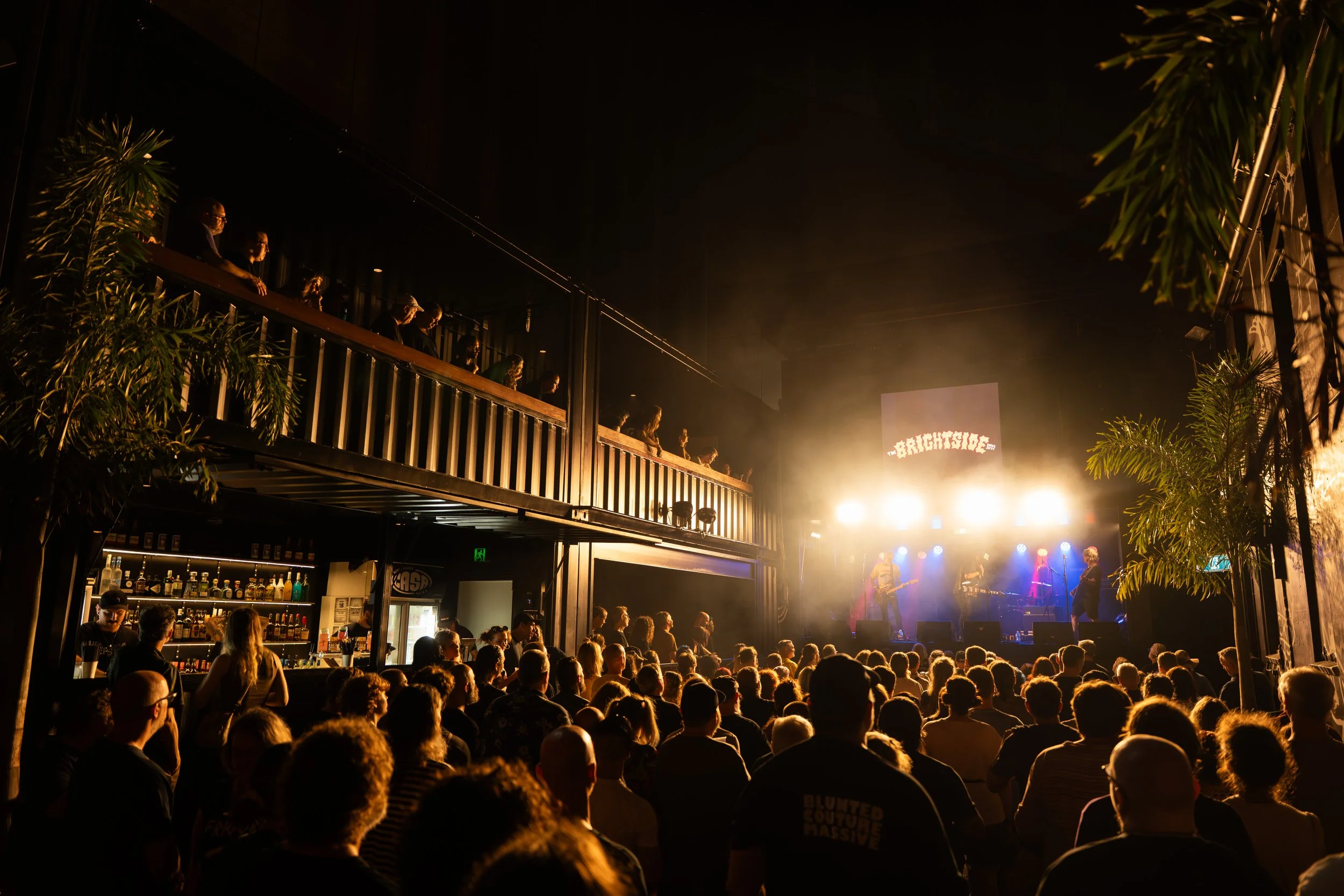 Indoor concert scene with a crowd watching a band perform on stage, illuminated by bright lights, with a second level balcony filled with spectators, and tropical plants on the sides.