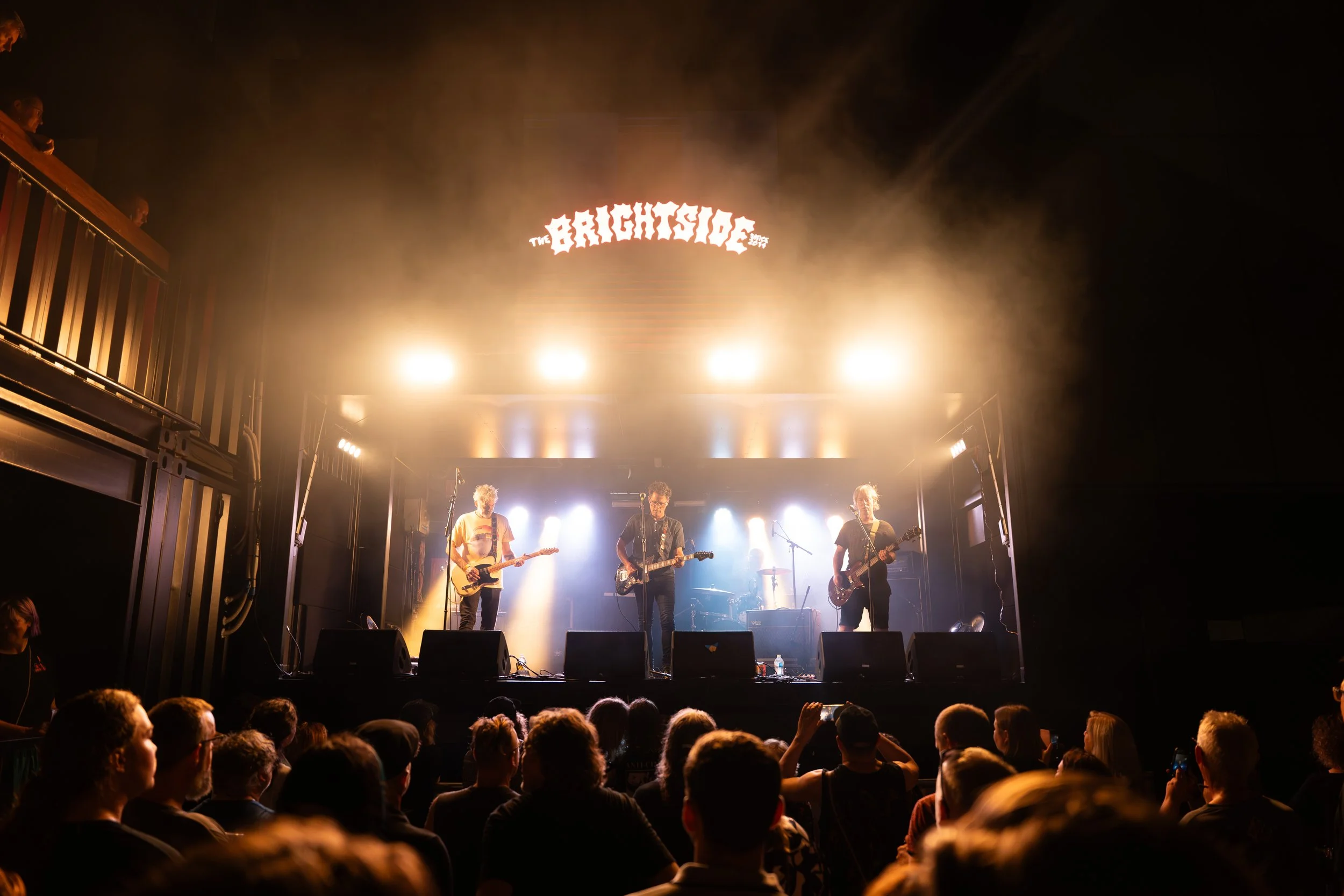 A band performs on stage at a concert with bright lights and smoke, with an audience watching.