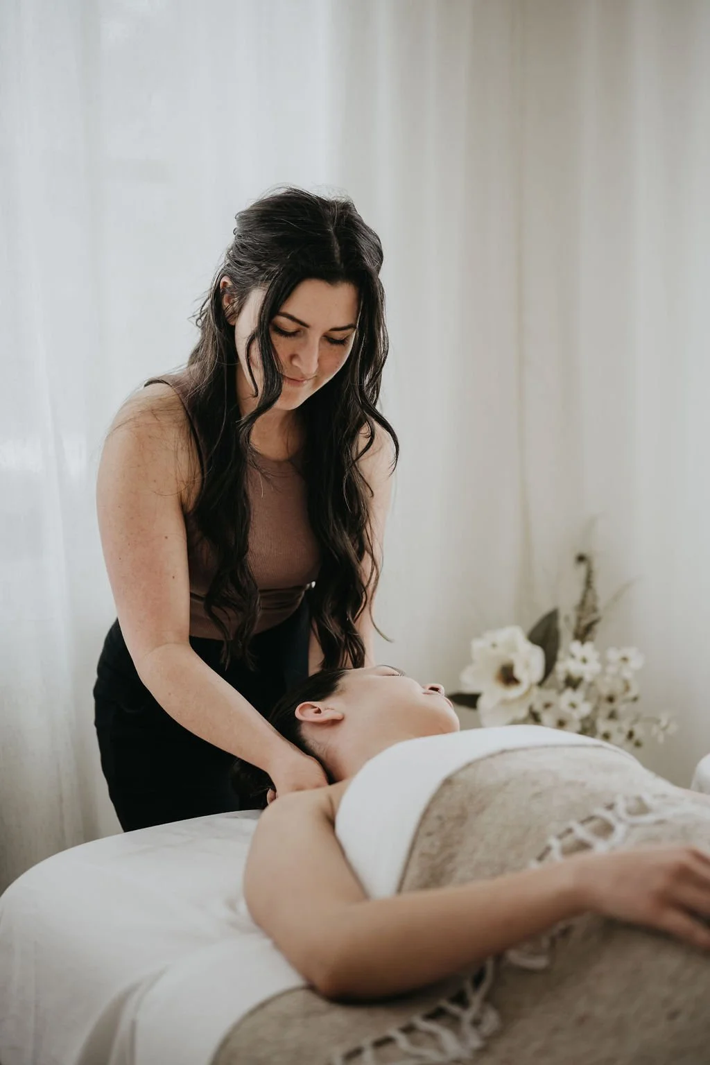 A woman is giving a massage to another woman lying on a massage table in a softly lit room with white curtains and floral decorations in the background.