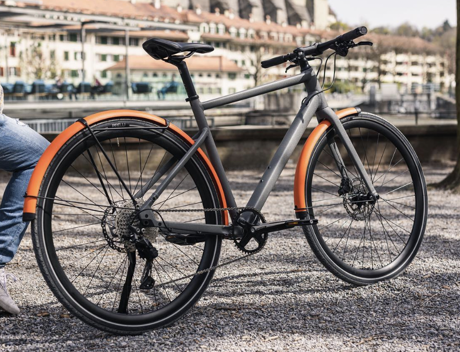 Gray and orange electric bicycle parked on gravel near water with buildings and trees in the background.