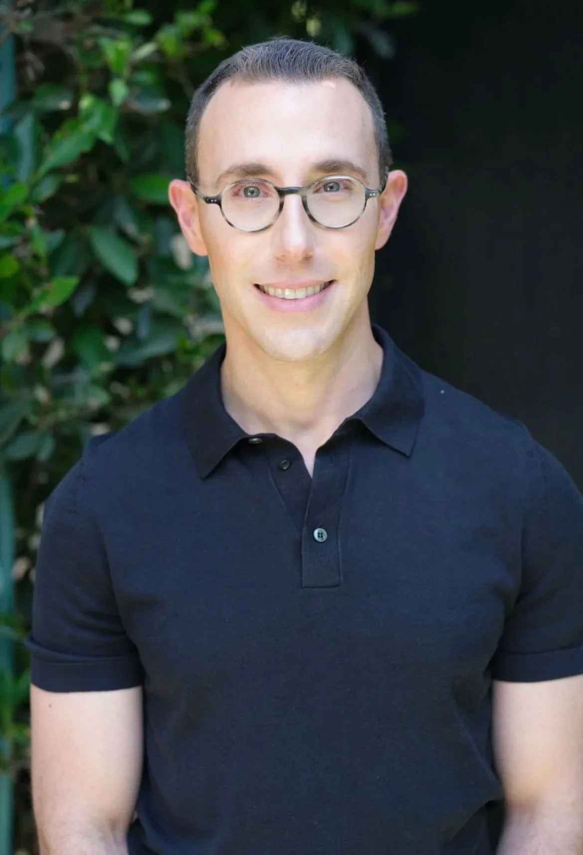 A smiling man with short brown hair, wearing glasses and a black polo shirt, standing outdoors with green foliage in the background.