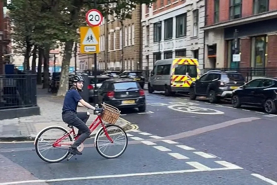A woman riding a red bicycle with a basket, at a city crosswalk with multiple parked cars and buildings in the background.