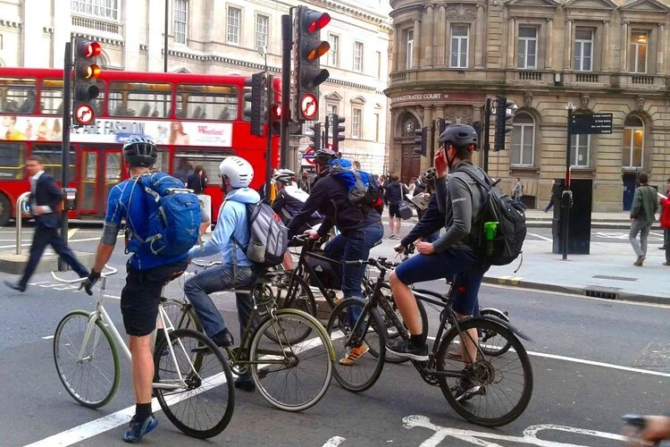 Group of cyclists waiting at a traffic light in an urban setting with historic buildings and a red double-decker bus in the background.